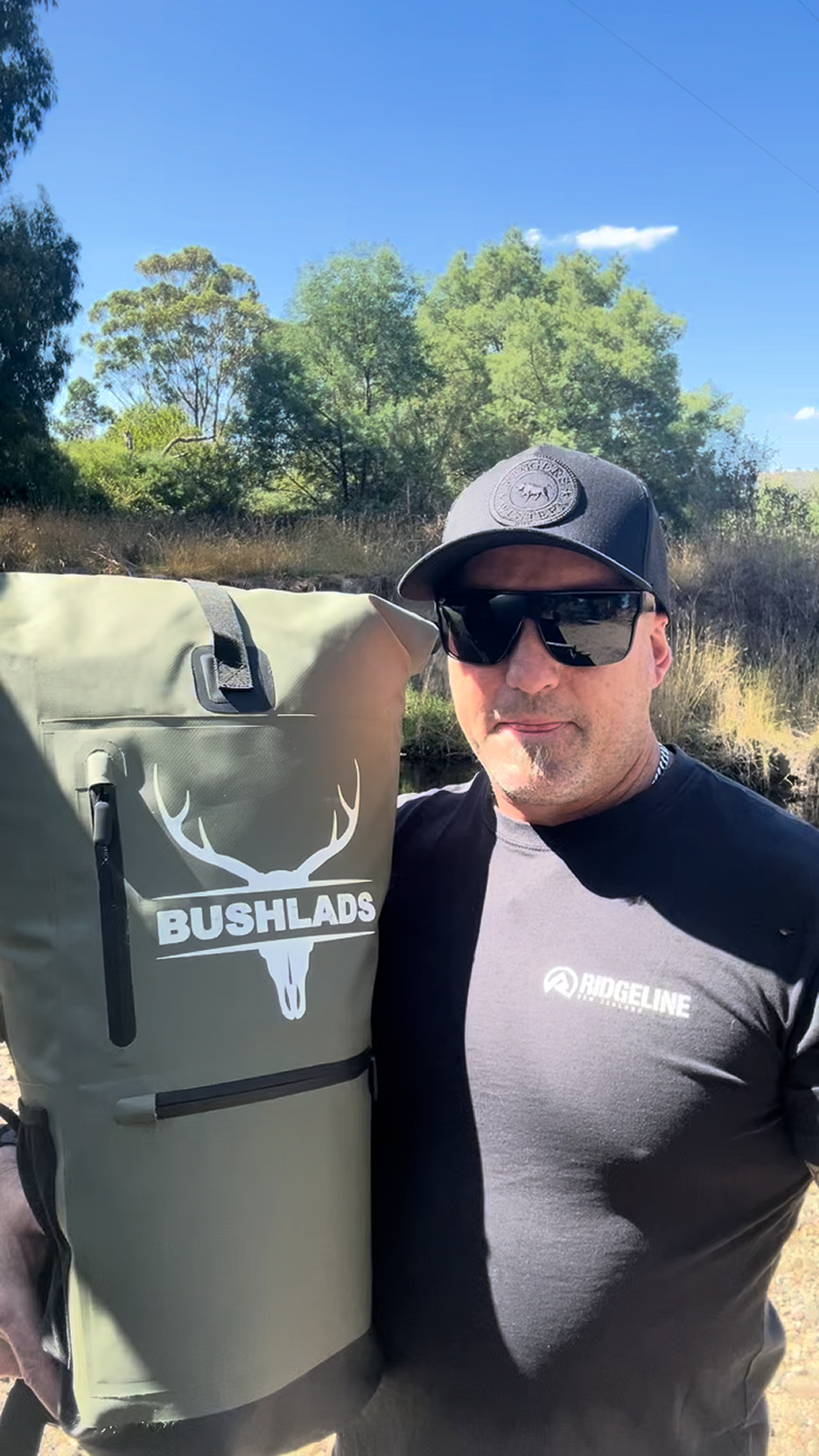 A man wearing a black cap and sunglasses holds a large olive green dry bag backpack with a white deer skull logo and the text 'BUSHLADS' on the front. The background shows trees and a blue sky outdoors.