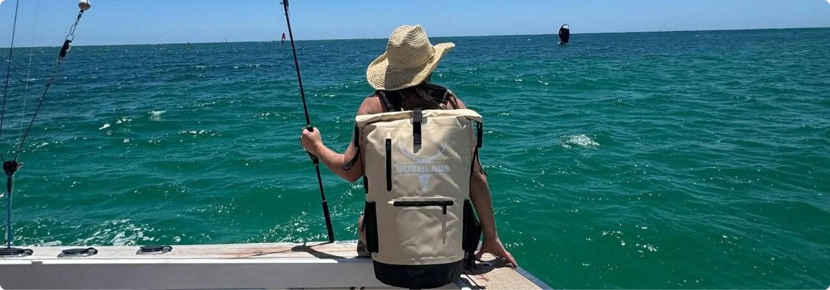 Angler in a straw hat sitting on a boat deck with a fishing rod, wearing the Bushlads Insulated Backpack in Sandy Beige over calm turquoise water