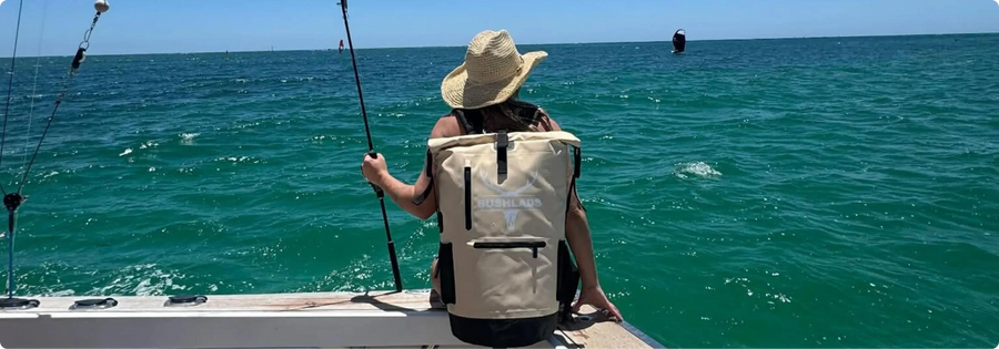 Angler in a straw hat sitting on a boat deck with a fishing rod, wearing the Bushlads Insulated Backpack in Sandy Beige over calm turquoise water